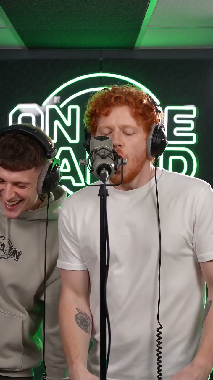 A red-haired man in a white T-shirt and headphones raps at the mic in the green booth, a man in a hoodie laughing beside him